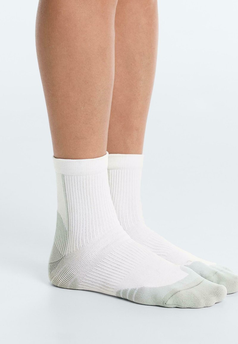 Beine mit weißen Knöchelsocken, die helle graue verstärkte Zehen- und Fersenbereiche haben, stehen auf einer weißen Oberfläche.