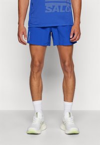 Blå sportshorts gjorda av lättviktsmaterial, med elastisk midja. Kombineras med vita sneakers och strumpor i ankellängd.