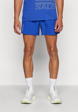Pantalones cortos deportivos azules hechos de una tela ligera, con una cinturilla elástica. Combinados con zapatillas blancas y calcetines de longitud media.