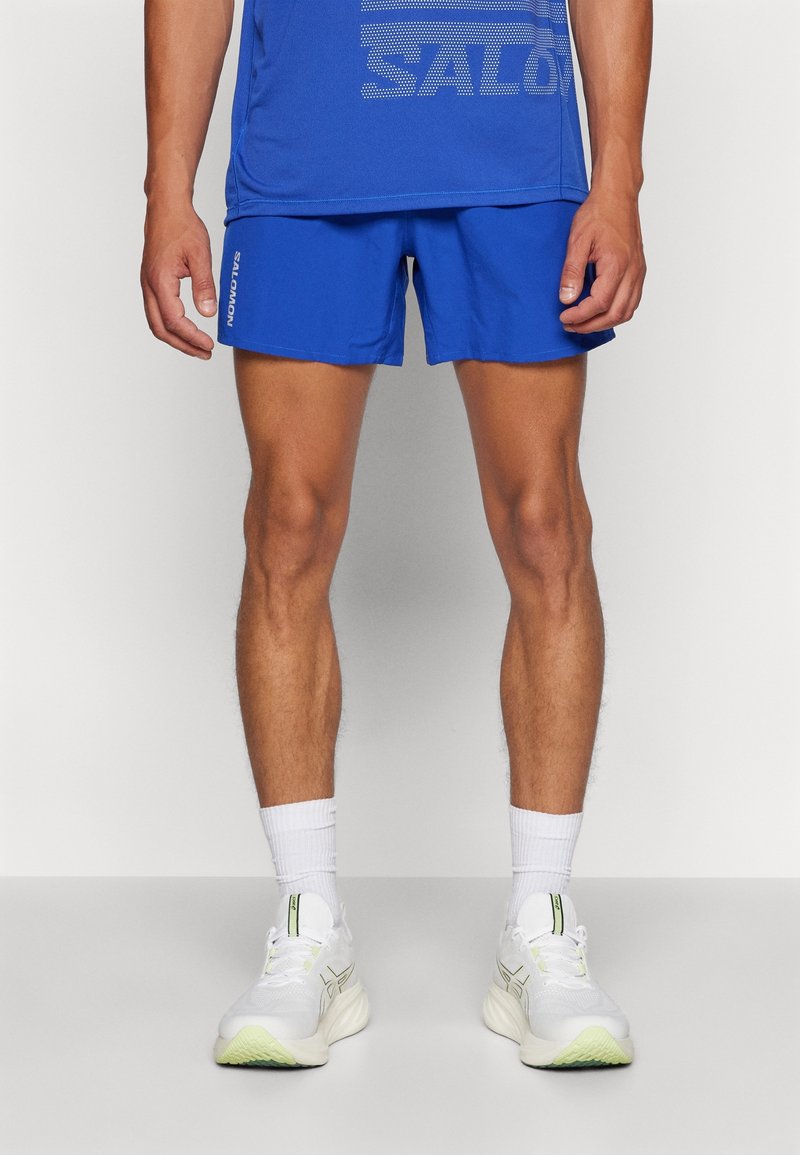 Blå sportshorts gjorda av lättviktsmaterial, med elastisk midja. Kombineras med vita sneakers och strumpor i ankellängd.
