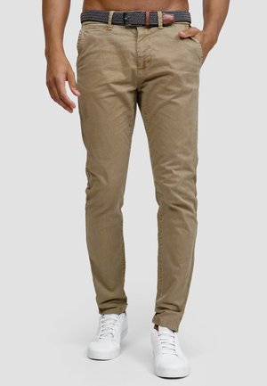 Mann trägt beige, eng anliegende Hosen mit einem schwarz-weißen gemusterten Gürtel und weißen Sneakern, steht mit einer Hand in der Tasche.