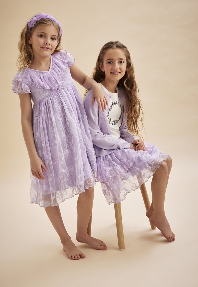 Två flickor i lavendelfärgade klänningar med spetsdetaljer. Den ena har en kortärmad klänning, medan den andra har en lagerkjol med blommönster.