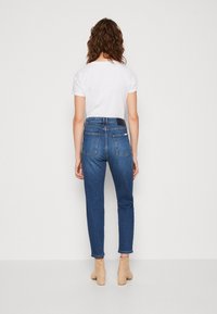 Jeans de denim azul con un diseño ajustado, estilo de cinco bolsillos y un dobladillo ligeramente corto. Combinados con una camiseta blanca lisa y botas de tacón cuadrado beige.