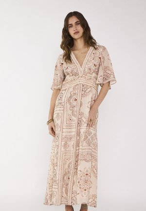 Femme aux cheveux ondulés portant une robe maxi crème avec des motifs floraux et géométriques couleur rouille, manches courtes et décolleté en V, debout calmement.