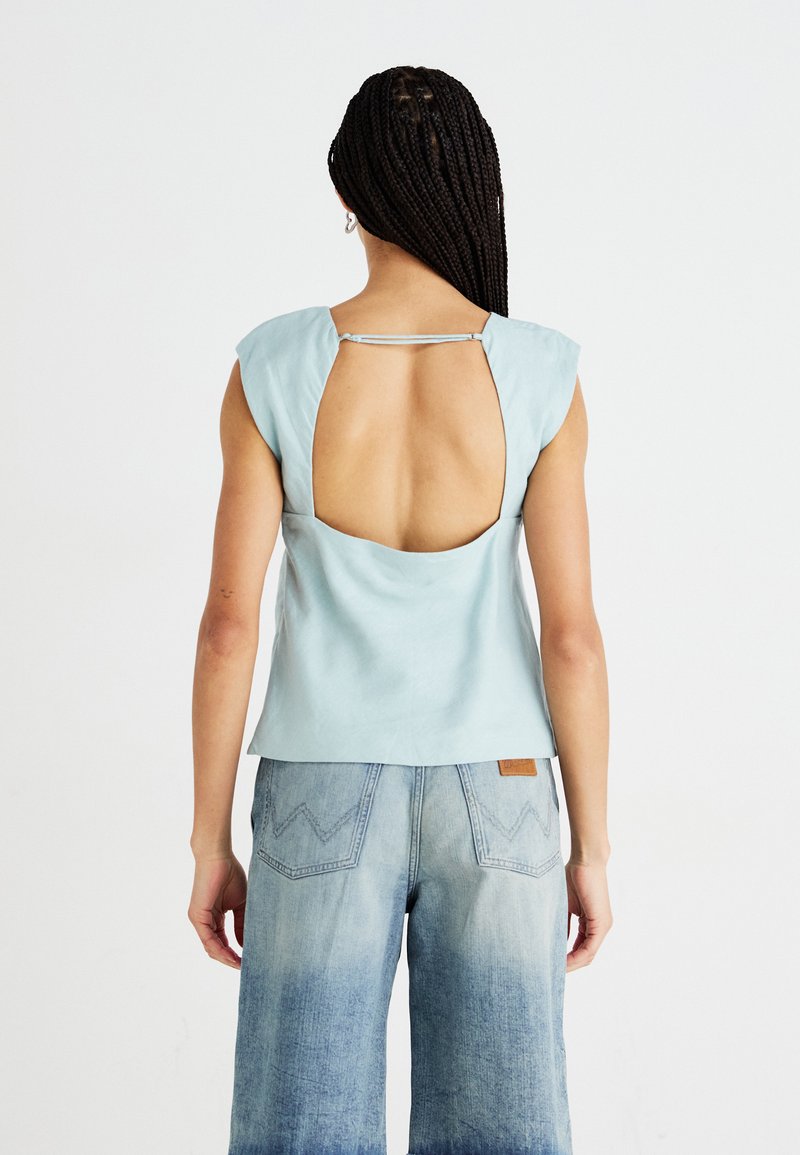 Haut sans manches bleu clair avec un design dos ouvert. Texture lisse, ajusté aux épaules, avec un petit détail de bretelle. Associé à un jean en denim.