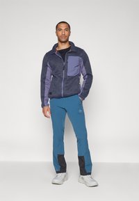 Veste en polaire bleu foncé avec des accents violets et une poche frontale, associée à un pantalon bleu avec des panneaux inférieurs noirs et des baskets grises.