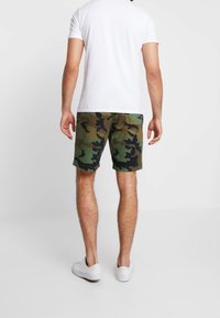 Shorts camouflage en tissu vert et marron, avec une coupe mi-cuisse, une poche arrière, associés à un t-shirt blanc uni et des baskets blanches.
