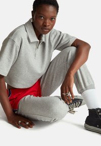 Šedé polo tričko s krátkými rukávy a polovičním zipem, kombinované se šedými tepláky s červenými detaily. Oblečení doplňují černé tenisky a prsteny.