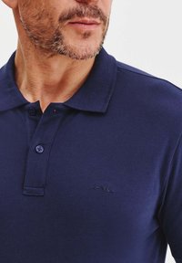 Granatowa koszulka polo z fakturowanej tkaniny, z krótkim kołnierzykiem, posiadająca dwa guziki i mały haftowany logo na piersi.