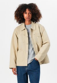 Beige jakke med krave, forlommer og trykknapper. Bæres over en grå skjorte og parret med blå jeans. Blød tekstur.