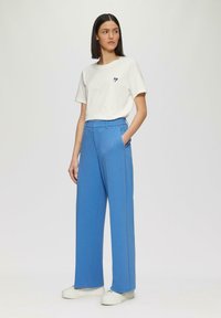 T-shirt bianco in cotone con piccolo dettaglio a cuore nero, abbinato a pantaloni larghi blu a vita alta e sneakers bianche.
