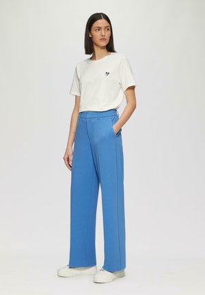 Weißes Baumwoll-T-Shirt mit kleinem schwarzen Herz-Detail, kombiniert mit hochgeschnittenen blauen weiten Hosen und weißen Sneakers.