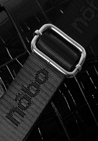 Matériau embossé croco noir avec une finition texturée. Doté d'une boucle en métal argenté et d'une sangle tressée ornée de "müabiö" en noir.