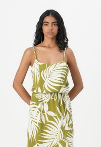 Robe d'été verte à imprimé tropical ornée de motifs de feuilles blanches, avec de fines bretelles spaghetti et une taille élastique pour un ajustement décontracté.