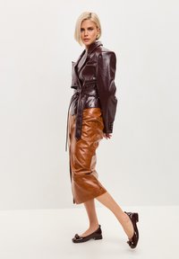 Veste en simili cuir bordeaux brillant avec taille ceinturée, associée à une jupe crayon en cuir marron et des talons noirs brillants avec des accents décoratifs.