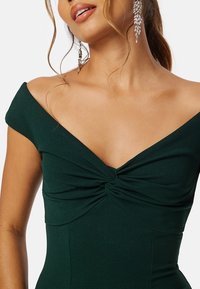 Abito verde senza spalline con dettaglio intrecciato al centro del busto. Tessuto liscio e aderente con una leggera lucentezza. Eleganti orecchini pendenti in argento.