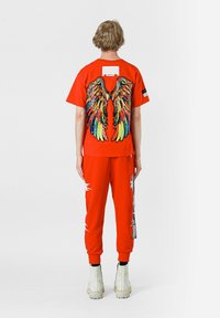 Camisa roja de manga corta con un diseño de alas de lentejuelas multicolores en la parte posterior, combinada con pantalones jogger rojos con detalles gráficos blancos y botas blancas.