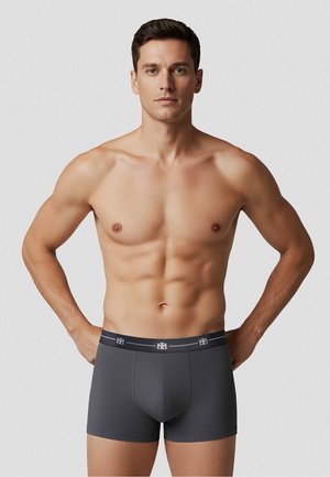 Muskuløs mand iført mørkegrå boxershorts, stående med hænderne på hoften mod en ensfarvet lys baggrund.