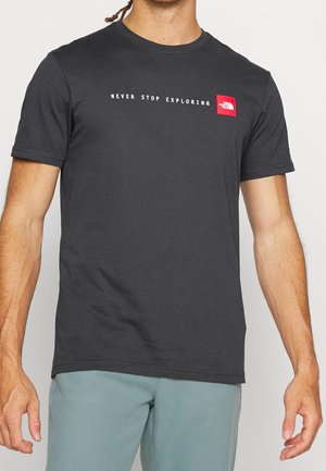 Mężczyzna noszący ciemnoszarą koszulkę z napisem "NEVER STOP EXPLORING" i czerwonym logo The North Face na piersi, w połączeniu z jasnoniebieskimi spodniami.