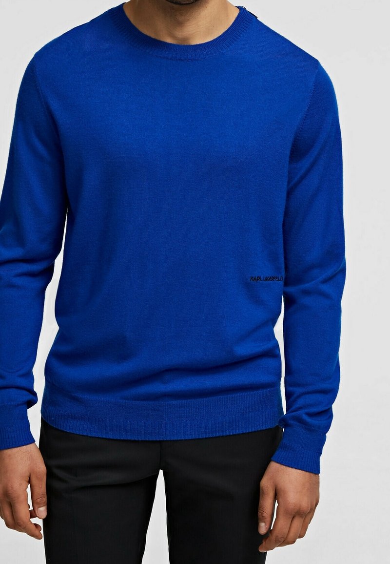 Blauer Strickpullover mit langen Ärmeln, Rundhalsausschnitt und geripptem Saum. Verfügt über ein dezentes Logo nahe der unteren Kante. Glatte Textur, leichtes Material.