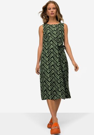 Un vestito verde senza maniche con un motivo a zig zag nero, caratterizzato da un dettaglio con fiocco in vita. Abbinato a sandali con zeppa arancioni.