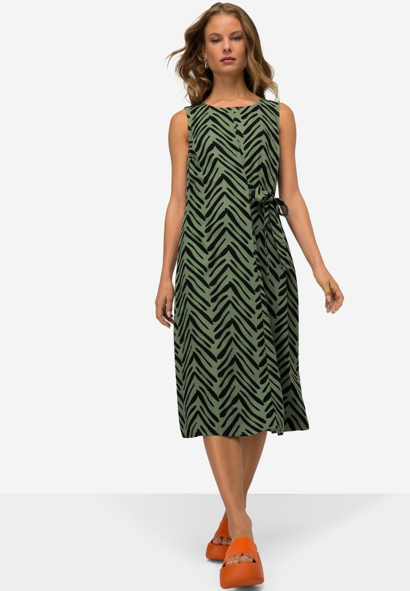 Ein grünes ärmelloses Kleid mit einem schwarzen Zickzackmuster, das mit einem Bindeband in der Taille ausgestattet ist. Kombiniert mit orangen Plateausandalen.