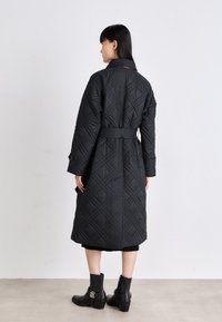 Μαύρο, καπιτονέ παλτό με ζώνη στη μέση, χαρακτηρισμένο από διαμαντένιο μοτίβο και μακριά μανίκια. Συνδυασμένο με μπότες μέχρι τον αστράγαλο, φορεμένο πάνω από μαύρη ενδυμασία.