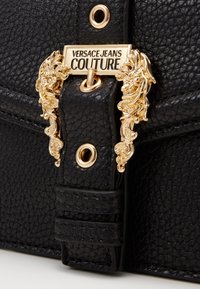 Svart texturerad läderbälte med utsmyckad guldsplitt med "Versace Jeans Couture"-logotyp och blomsterdekorationer.