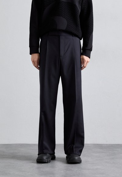 Pantaloni neri su misura con un design a gamba dritta, caratterizzati da una texture liscia e pieghe sottili, abbinati a scarpe da ginnastica nere.