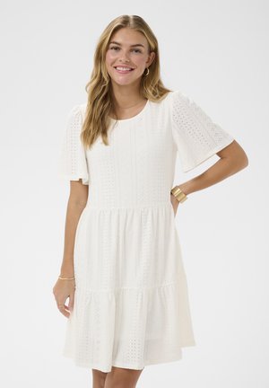 Uśmiechnięta kobieta o długich blond włosach, ubrana w białą sukienkę z krótkim rękawem zdobioną ażurowym wzorem oraz złotą biżuterią, stoi z jedną ręką na biodrze.