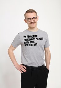 Šedé bavlněné tričko s tučným černým textem "MÝM NEJOBLÍBENĚJŠÍM DĚTSKÝM VZPOMÍNKOU JE, ŽE NEBOLÍ ZÁDA." Nosí se s černými kalhotami.