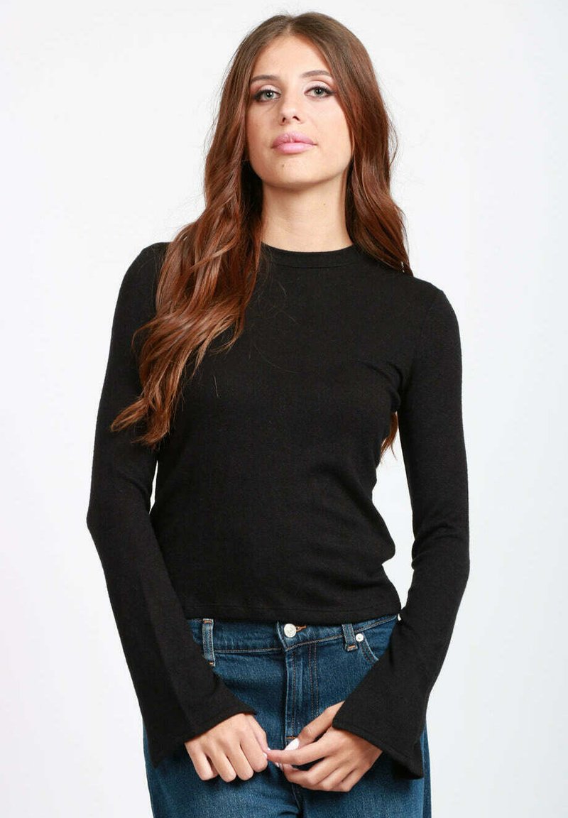 Donna con i capelli lunghi e marroni, vestita con una maglietta nera aderente a maniche lunghe e jeans blu, in piedi contro uno sfondo bianco semplice.