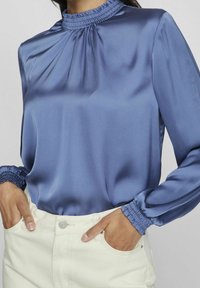 Satinbluse mit langen Ärmeln in Hellblau, mit gerafftem Rundhalsausschnitt und gepufften Manschetten. Kombiniert mit weißen, hochtaillierten Hosen.