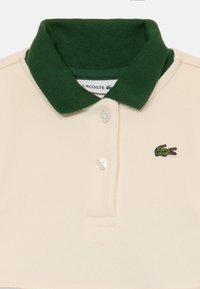 Polo crème avec col vert foncé, deux boutons blancs et petit logo crocodile vert sur la poitrine gauche.