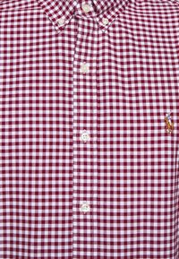 Red and white gingham button-up shirt, featuring a classic collar, multiple buttons, and a small embroidered logo on the chest.