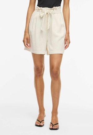 Kvinne iført beige shorts med høy midje med et stoffbelte bundet i en sløyfe og svart ermeløs topp, står foran en hvit bakgrunn.