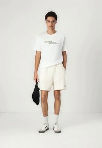 Junger Mann steht, trägt ein weißes "Jack & Jones" T-Shirt, cremefarbene Shorts, weiße Socken, Turnschuhe und hält eine schwarze Umhängetasche in der linken Hand.