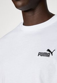 Valge puuvillane T-särk ümmarguse kaelusega, millel on must tikanditud Puma logo vasakul rinnal. Pehme tekstuur, klassikaline lõige.