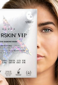 Emballage de masque facial en argent avec un design étoile holographique, présentant du texte noir et une icône de masque blanche, tenu partiellement devant un visage.