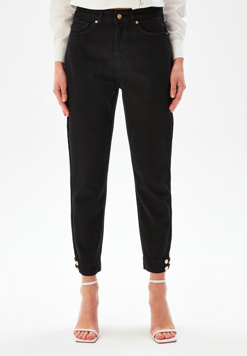 Personne portant un jean noir taille haute avec des boutons dorés à la cheville, un chemisier blanc et des sandales à talon blanches, debout devant un fond uni.