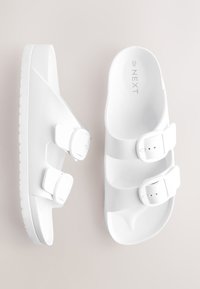 Sandales blanches à enfiler avec trois boucles réglables, fabriquées en matière plastique lisse, dotées d'une semelle texturée pour une meilleure adhérence.