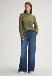 Maglione a maglia verde oliva con vestibilità rilassata e collo a costine, abbinato a jeans blu in denim a gamba larga e sneakers bianche.
