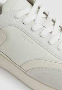 Sneaker bianco con una combinazione di pelle liscia e suede, caratterizzata da accenti testurizzati, forma strutturata e lacci bianchi. Logo stampato sul lato.
