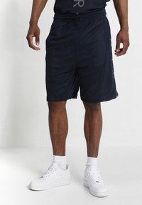 Man som bär svarta nät-idrottsbyxor, vita ankelsockor och vita Nike-sneakers, stående mot en plain vit bakgrund.