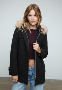 Čierny kabát z vlnenej zmesi s umelou kožušinou na kapucni, predným zipsom a dvoma veľkými náplecnými vreckami, nosený cez tmavý top a svetlé džínsy.