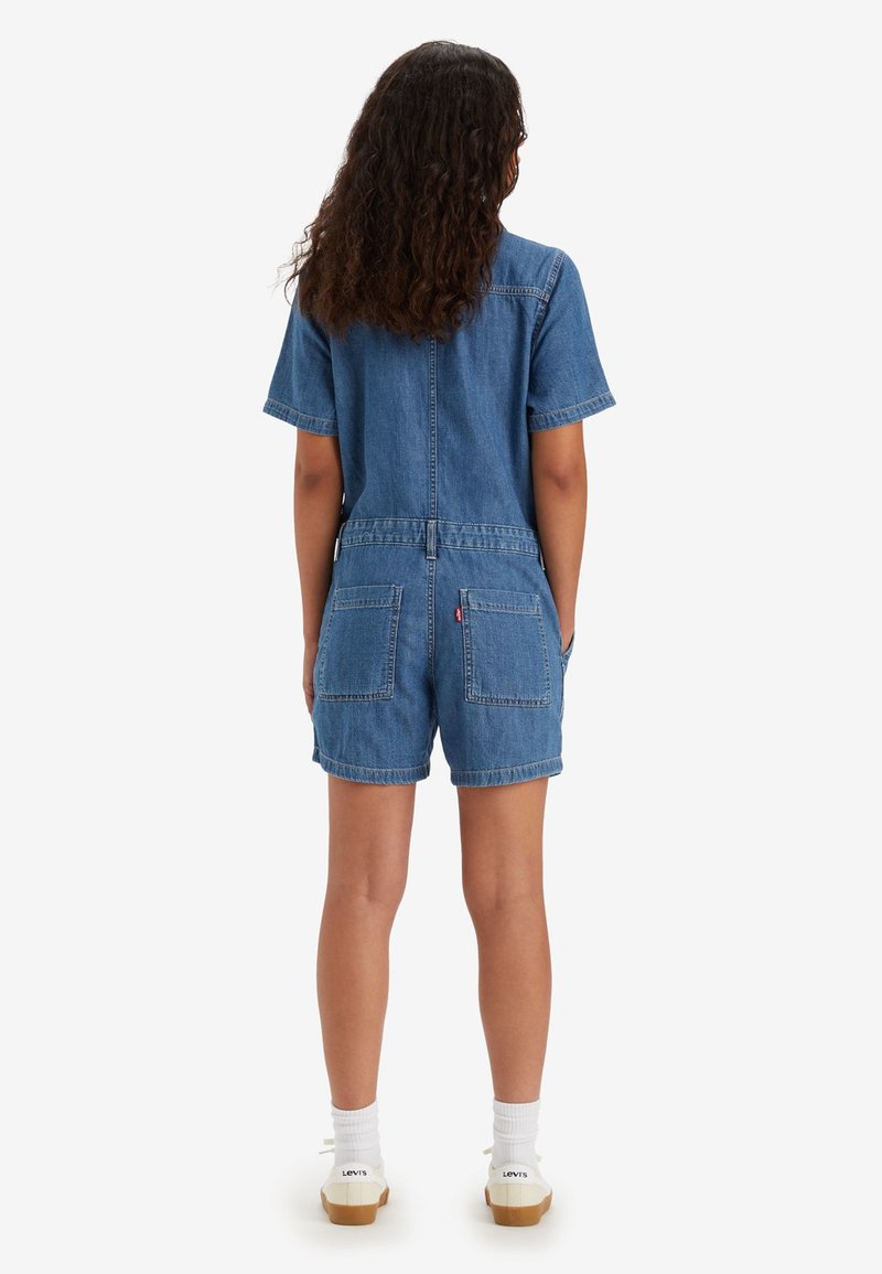 Jumpsuit en denim bleu clair, à manches courtes, avec deux poches arrières et une taille cintrée. Tissu texturé avec des détails de couture visibles.