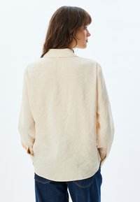 Licht beige langwerpige blouse met een relaxte pasvorm, zachte textuur, klassieke kraag en ronde zoom. Draagt over een blauwe spijkerbroek.