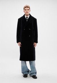 Μαύρο διπλόχρωμο παλτό από μαλλί με γιακά σε σάλι και κίτρινα κουμπιά, σε συνδυασμό με ανοιχτό μπλε φαρδύ τζιν και μαύρα παπούτσια.