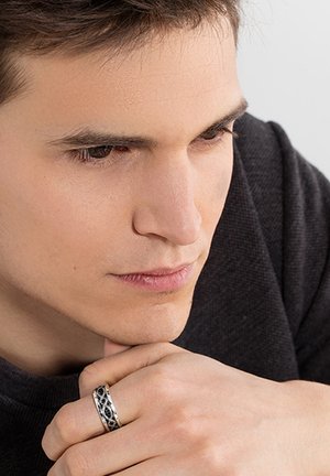 Jeune homme reposant son menton sur sa main, portant une bague noire texturée à son majeur, regardant vers le bas dans un cadre neutre.