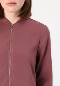 Burgundowa kurtka bomber z zamkiem błyskawicznym, okrągłym kołnierzem, dopasowanymi rękawami i gładką, elastyczną strukturą materiału.